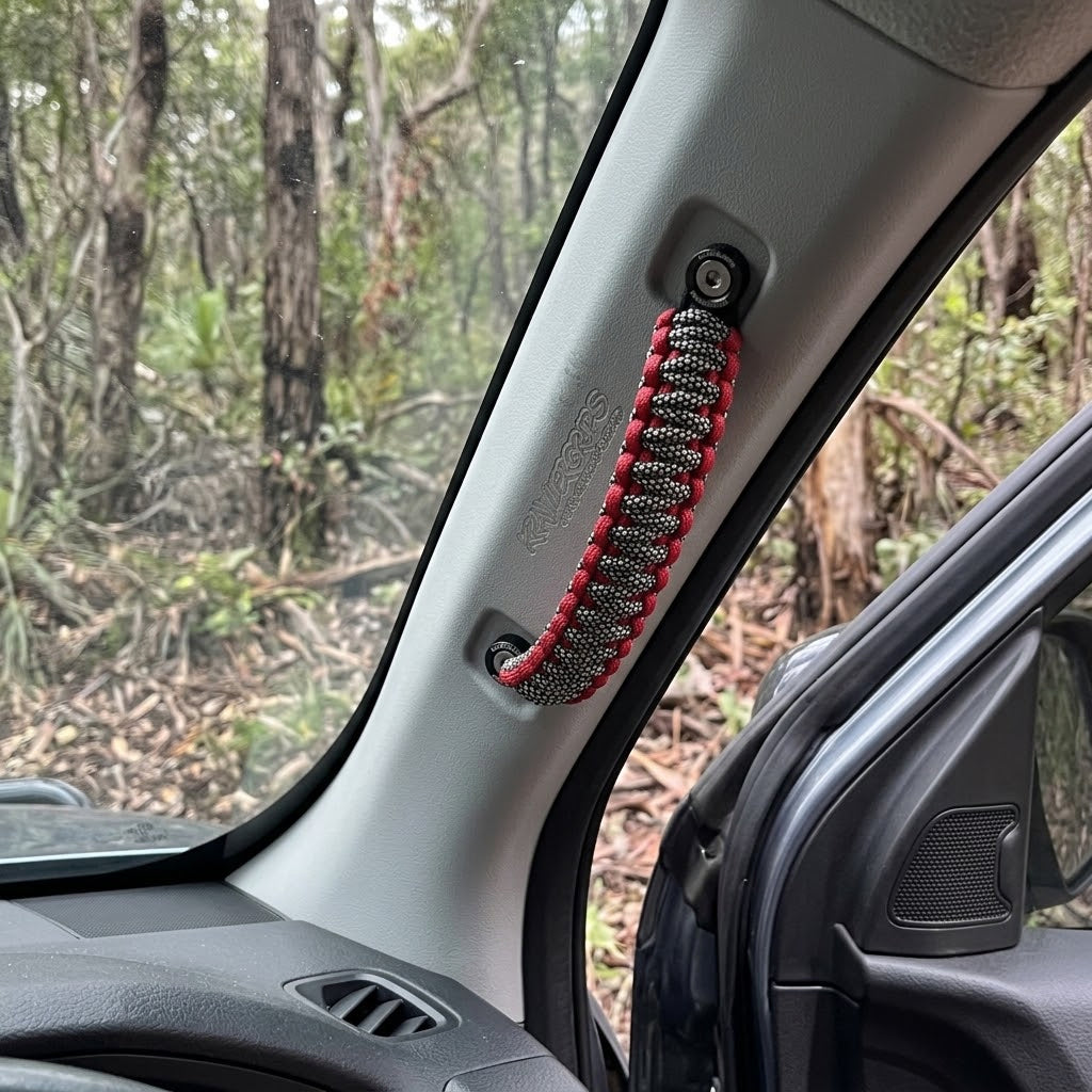 A red and black paracord car window grab handle mounted on a gray vehicle interior pillar, with a forest background visible through the window.