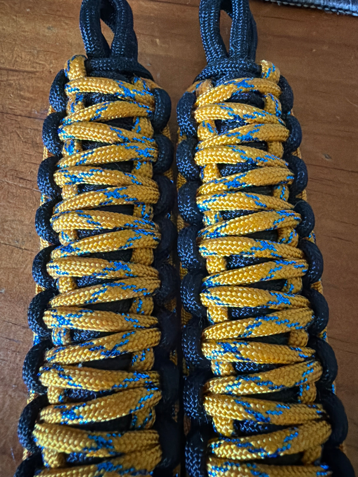 Two handmade paracord wrist wraps or tool wraps made from yellow and blue paracord with black braided edging, laid flat on a wooden surface.